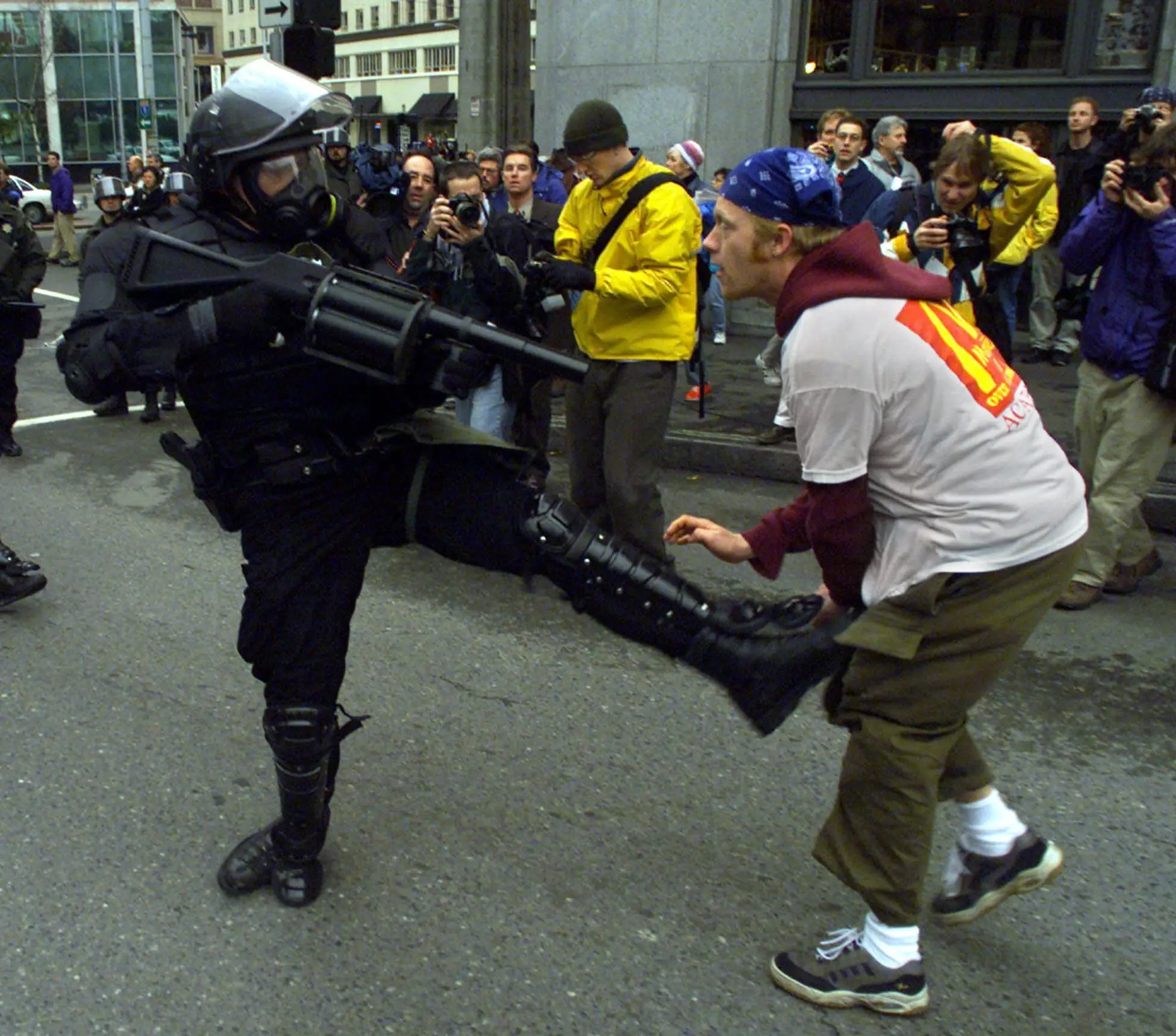 A heavily-armored officer points a large gun at a protester while kicking them.