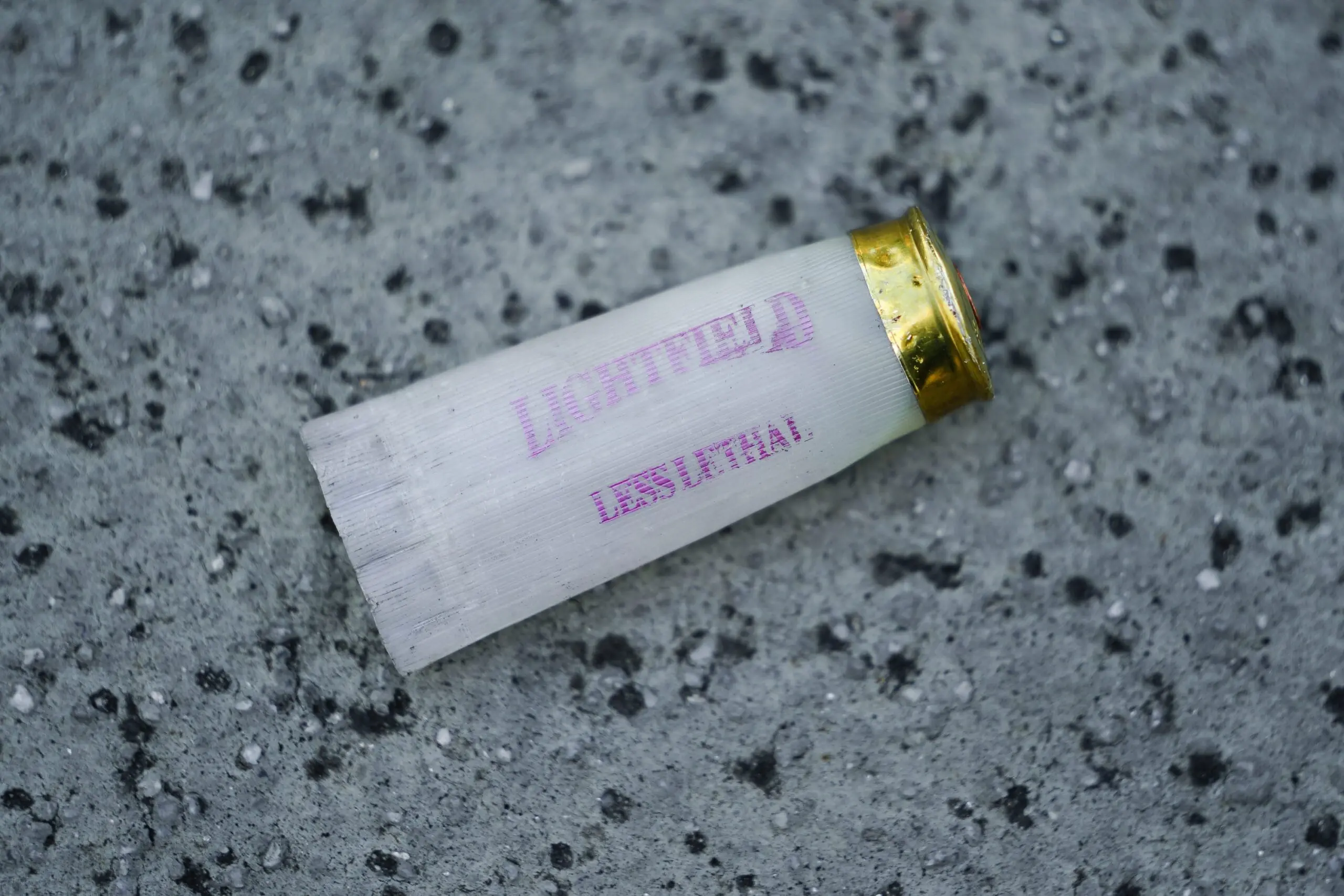 A shotgun shell with the words "less lethal" on it is crushed on the ground.