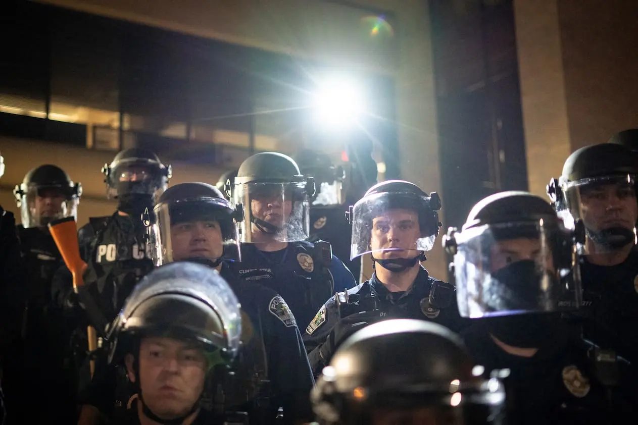 Rows of police officers in heavy gear face forward and shine a light towards the camera.