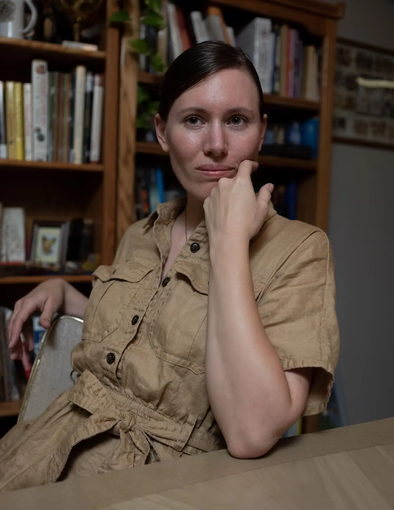 A woman sits in front of a bookshelf with her hand holding up her chin.
