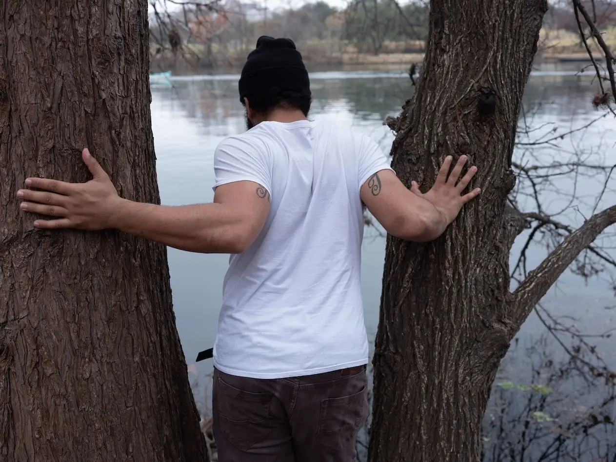 A man stands between, and holding, two trees, facing away from the camera and towards the water.